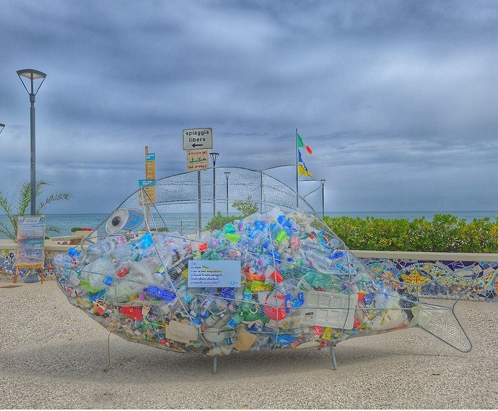 Colourful picture of a fish-shaped net with recyclable waste inside and a seafront view behind it. The sign above reads “Spiaggia libera” (free beach).