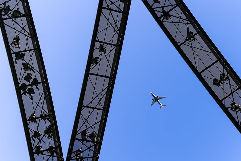 View from underneath and airplane flying in a clear blue sky between pedestrians visible on three transparent bridges in a W-like formation.