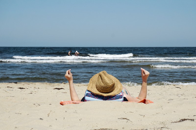 Picture of a person lying in the sand with hands up and wearing a straw hat, looking at the beach where there are two people standing among the waves.