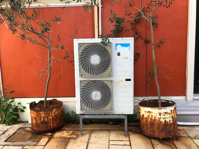 Picture of a double heat pump on legs, between two tree plants in rusted pots in a yard with an orange wall as background.