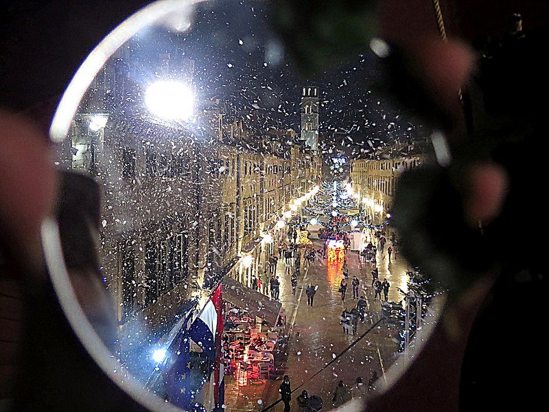 Picture viewed through a lens of a city pedestrian street with buildings on either side, a French flag visible on the left, and blurred fingers holding the lens.
