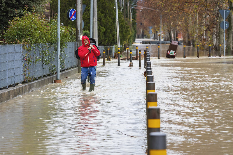 Picture of a flooded street – both pavement and road – with a man with a red hoodie seen walking on the left with water up to his knees.