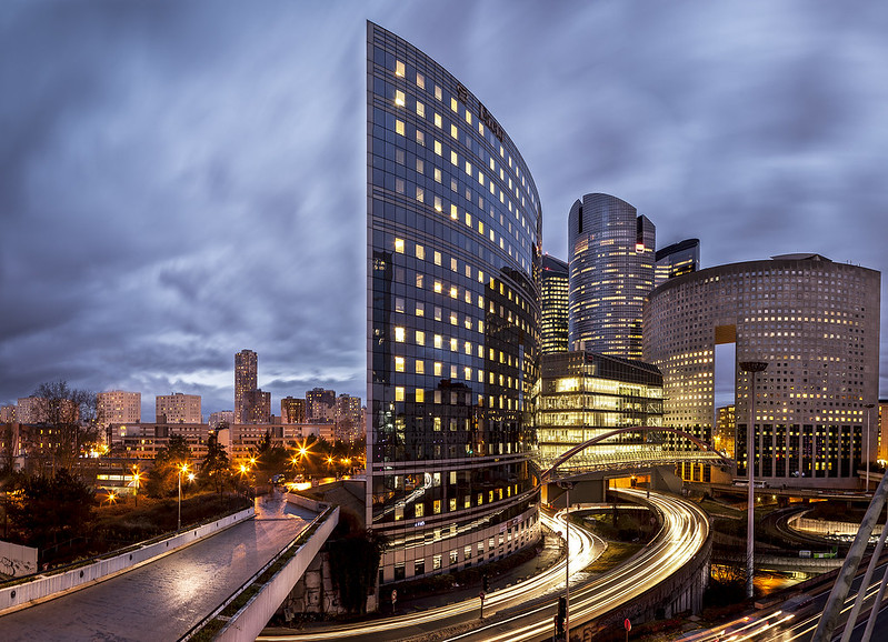 Picture of business area (La Defense, Paris) at dusk in a cloudy sky with lit street lamps and building lights visible.