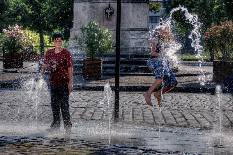 Picture of two young children playing in a water fountain in a town square with flowers at the back; one is looking at the camera, while the other is jumping in the water.