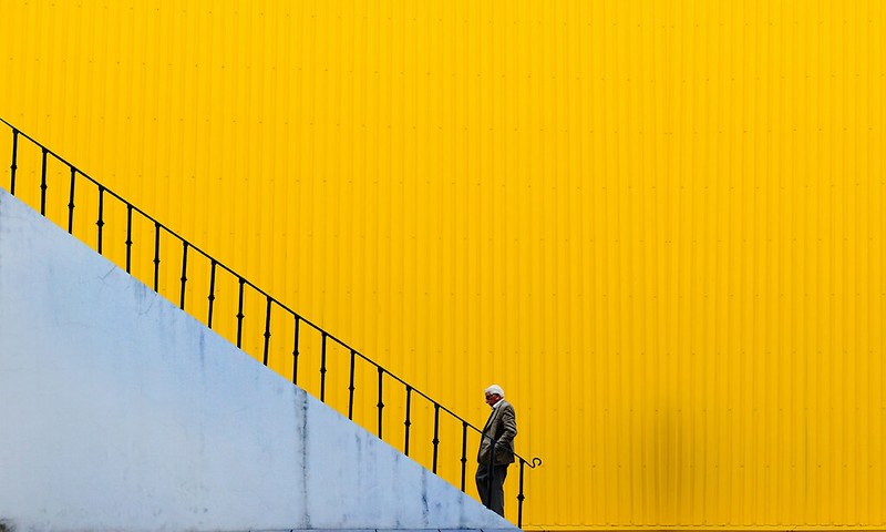 Profile image of an old man standing at the bottom of a staircase with a black railing and white bottom on a yellow background.