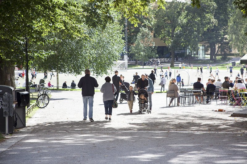 Image of people in a park on a sunny day, with trees visible on the left and in the background, and people walking or sitting at a café on the right.