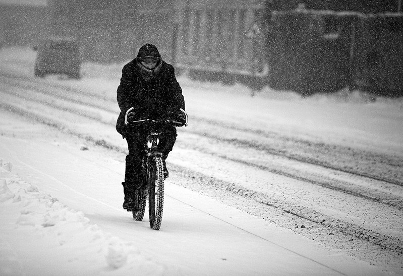 Black and white picture of a man riding a bicycle on a snow-covered road with a car visible in the snow-blurred background.