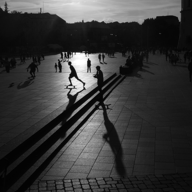 Black and white image of people in a pavement-tiled city square with the sun’s reflection visible at the centre.