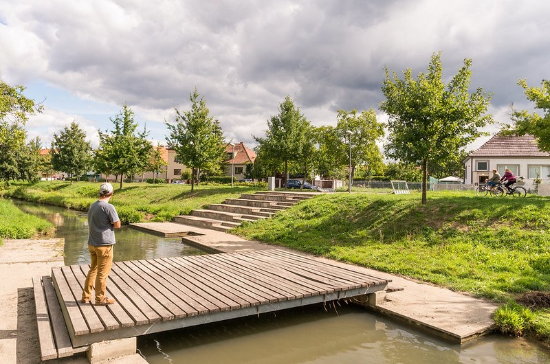 Picture of a person standing on a wooden bridge over a river and gazing into the background, with trees lining the river bed, a house and two cyclists visible on the right.