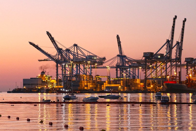 Picture of a container terminal at a port during dusk with a few small boats in the forefront in still water.