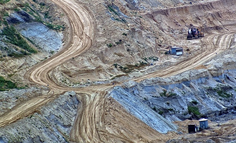 Picture of a mining quarry from a distance, a path is visible at the centre, as well as a bulldozer on the top right and a small container on the bottom right.