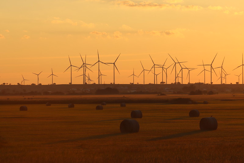 Picture of energy windmills in tandem in a yellow-orange background with a darker field with haybales in the forefront.