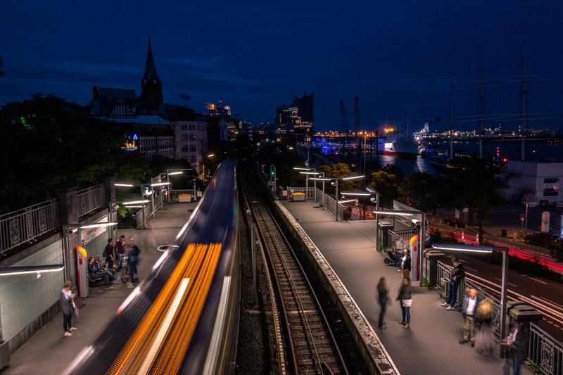 Picture of a railway station at night with the tracks in the centre and passengers waiting on the platforms on either side, and the city and port at the background.