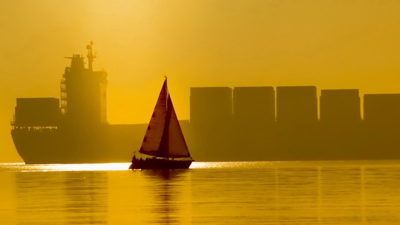 Picture of an orange sailboat in calm waters with the shadow of a large ship container visible in a yellow background.
