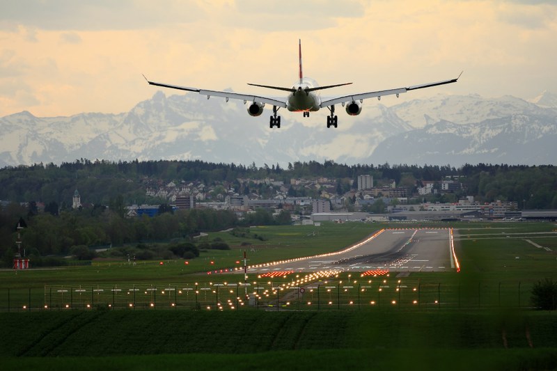 Picture of a plane landing on a green runway with the city in the background and the snowy hills in the distance.